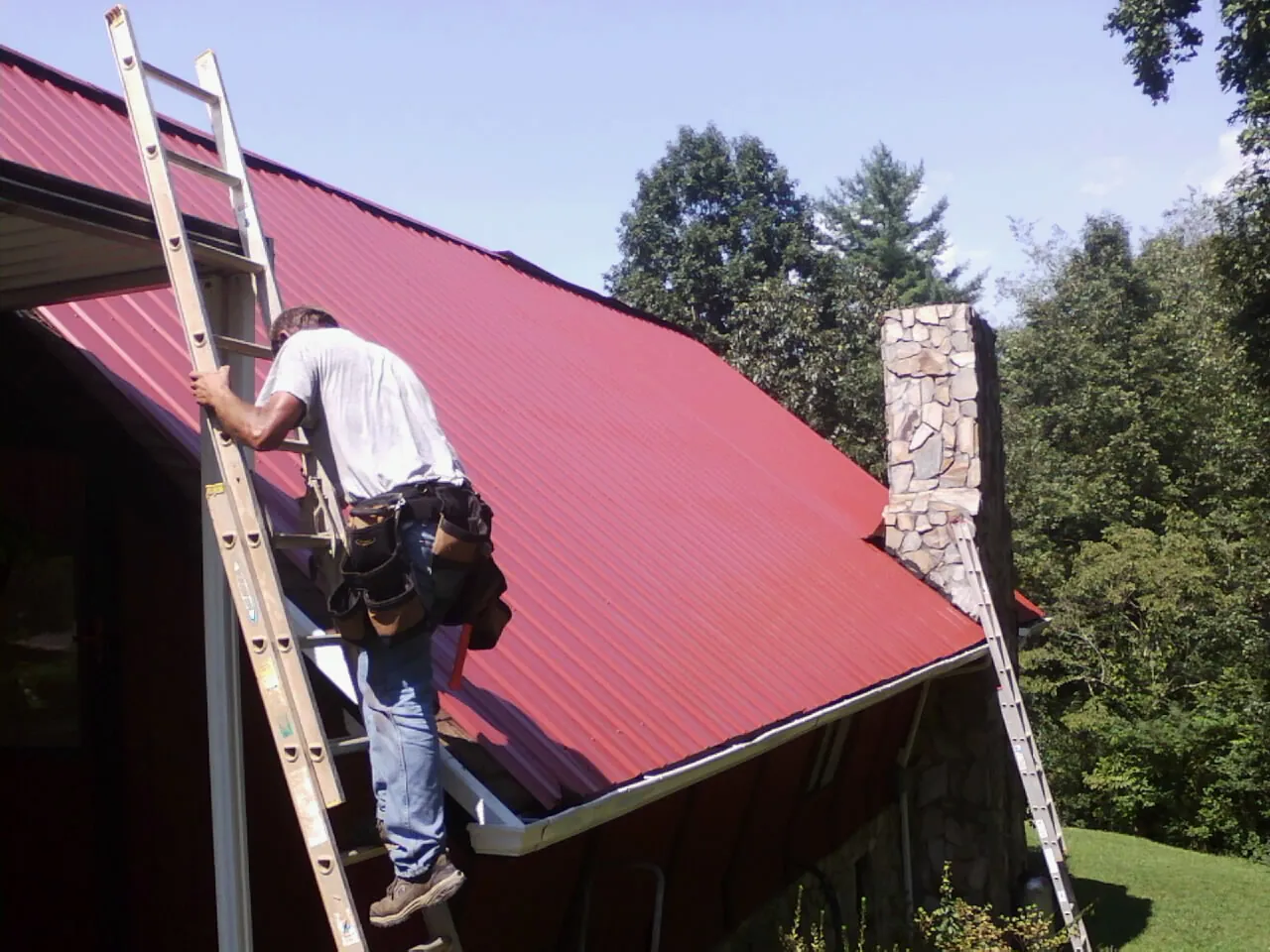 Roofer on a ladder beside a red metal roof and stone chimney — Soffit & Fascia Repair by Roofing Brothers in Gilbertsville
